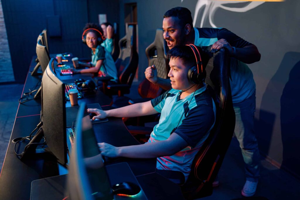 A gaming team competes in an esports tournament, with a coach guiding a player. All wear headsets and matching jerseys.