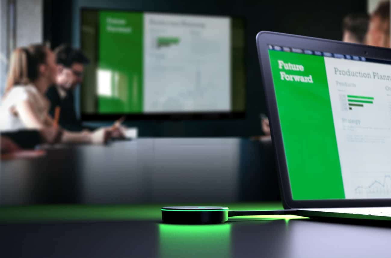 A Crestron conference room with a laptop showing Future Forward presentation, background screen with graphs, people discussing, glowing green device.