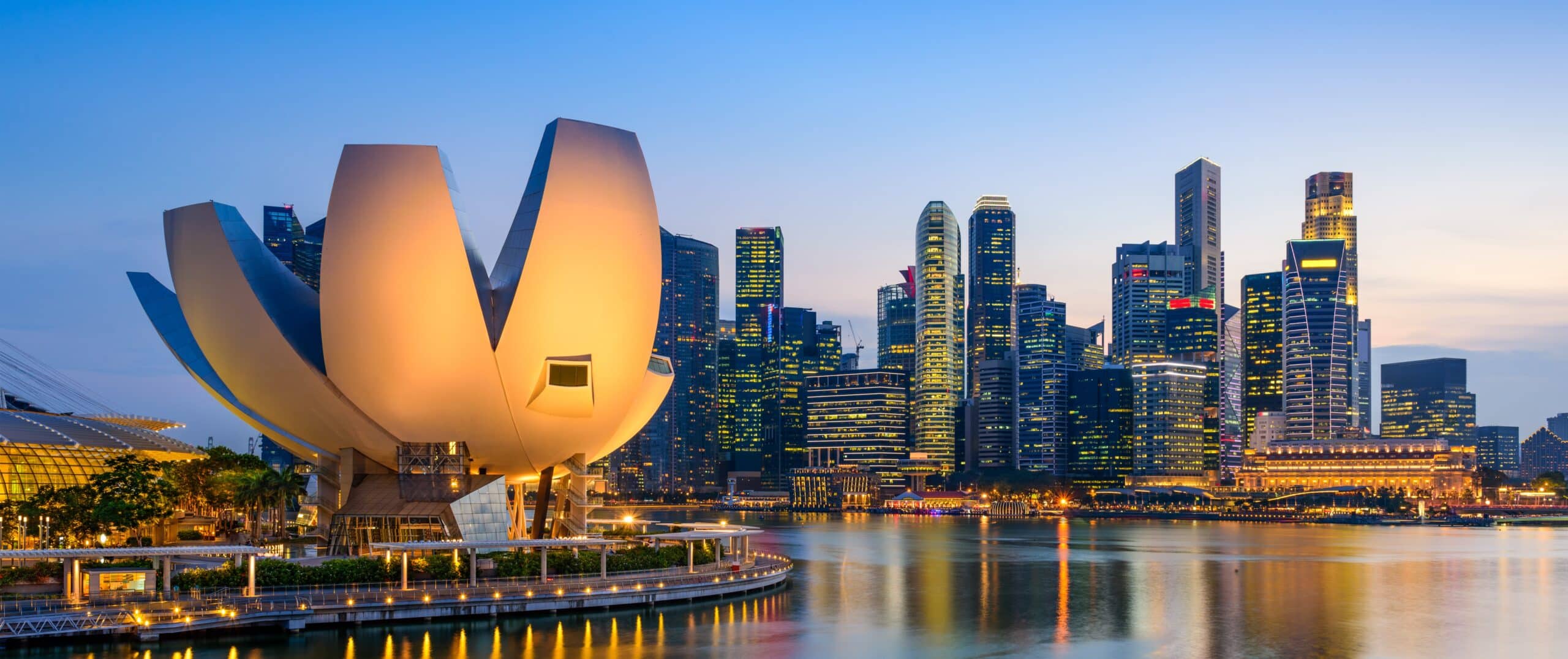 Singapore skyline at dusk with the lit-up ArtScience Museum in the foreground and glowing skyscrapers against a clear sky.