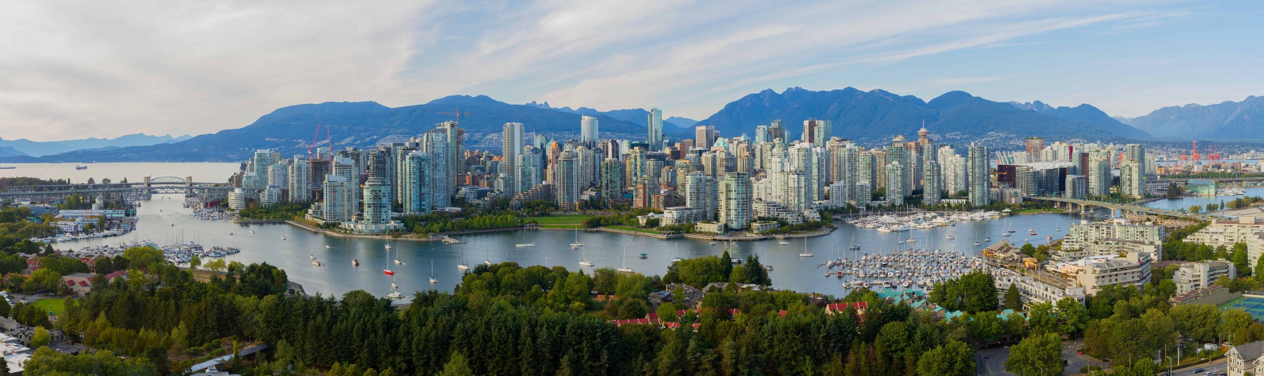 City skyline with tall buildings, water, boats in marina; mountains and partly cloudy sky backdrop; green trees in the foreground.
