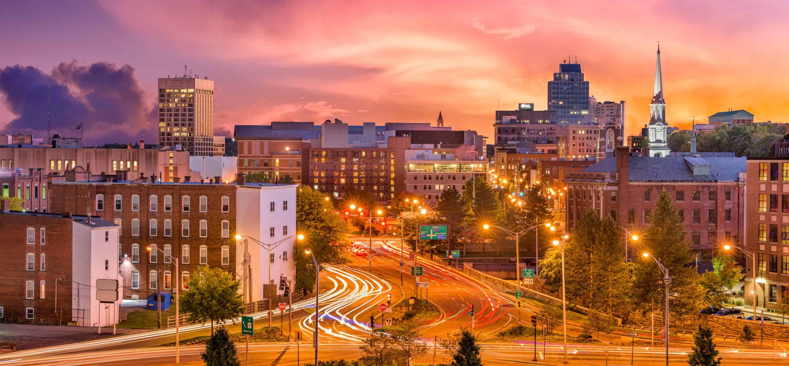 Vibrant cityscape at sunset: colorful clouds, lit streets, vehicle light trails, skyline buildings & church steeple.