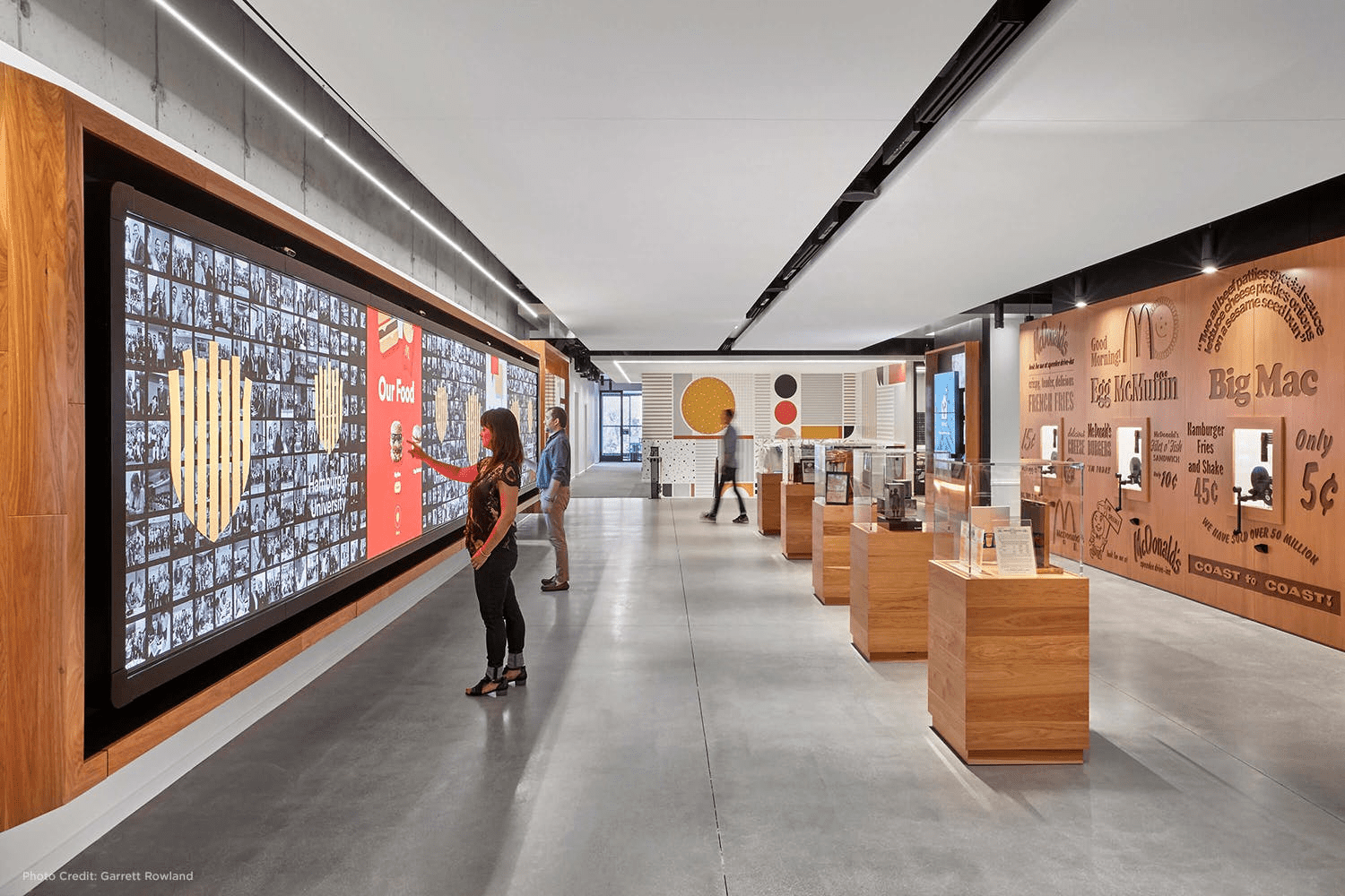 Modern museum exhibit with interactive displays, large screens, sleek design, wooden & metallic accents. Visitors engaging actively.