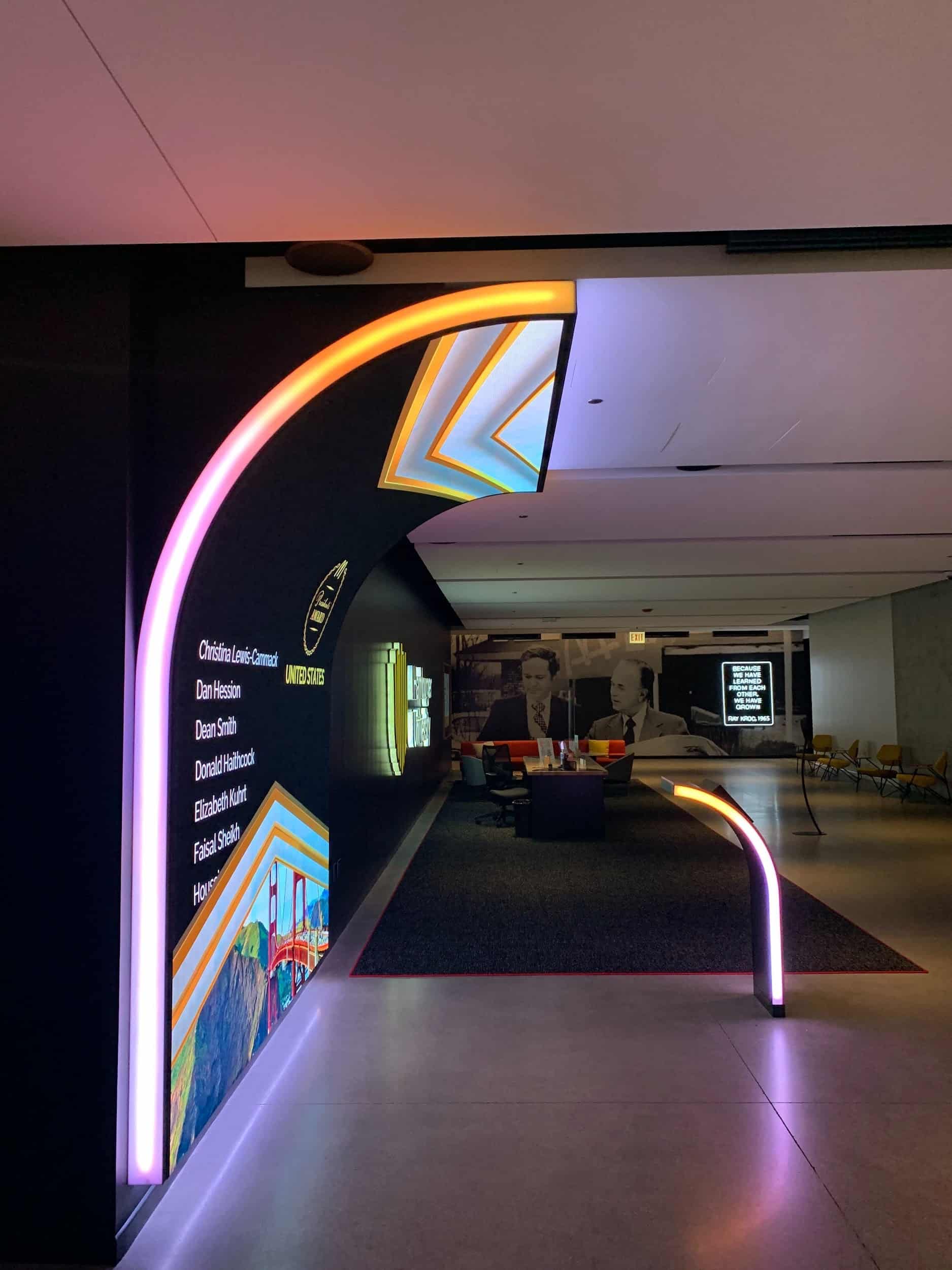 Modern hallway with neon arch, names on the left wall; vibrant designs. Background has colorful seating and a B&W mural.