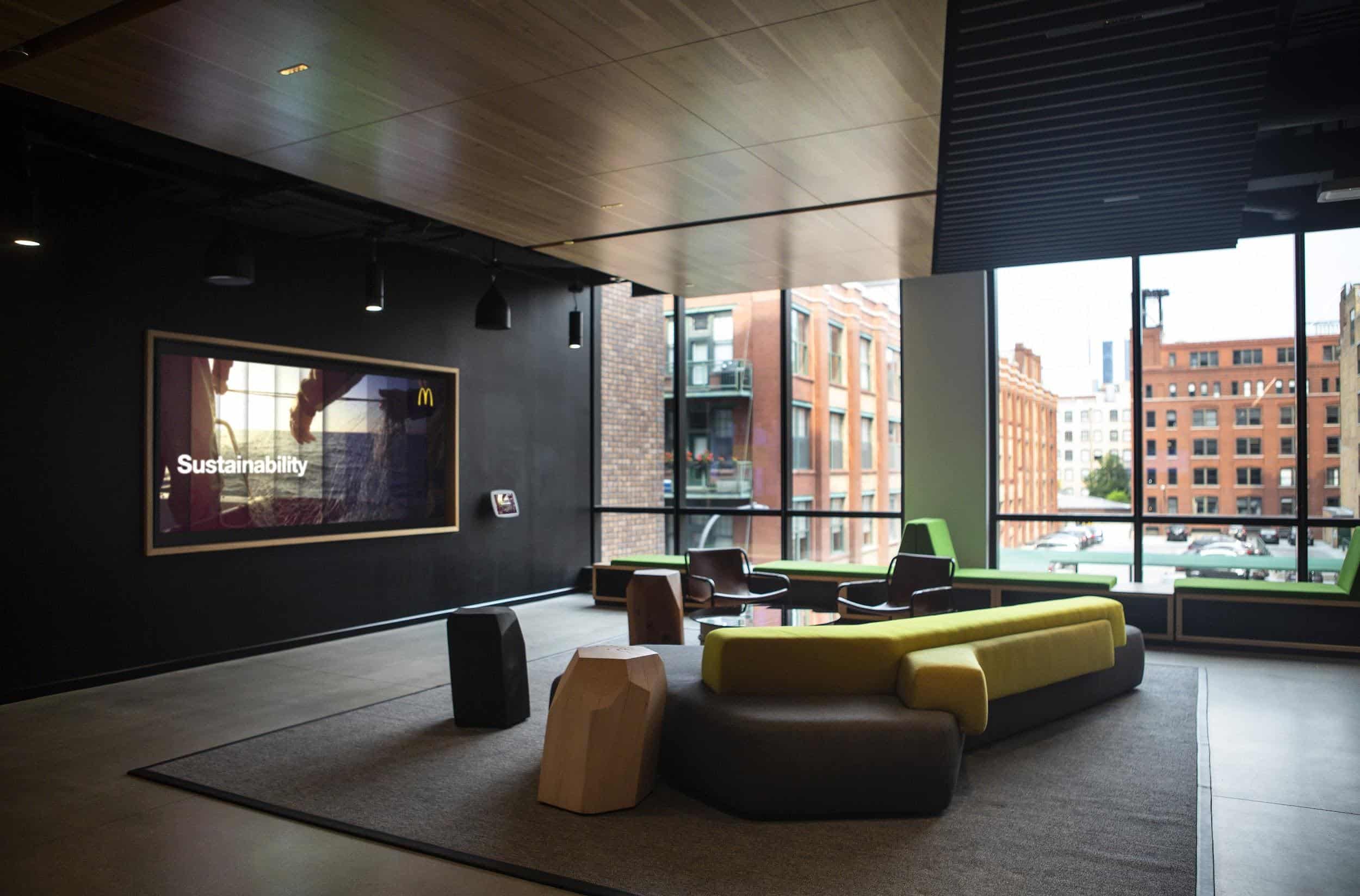 Modern office lounge: green/yellow sofa, dark chairs, city-view window; wall screen reads Sustainability.