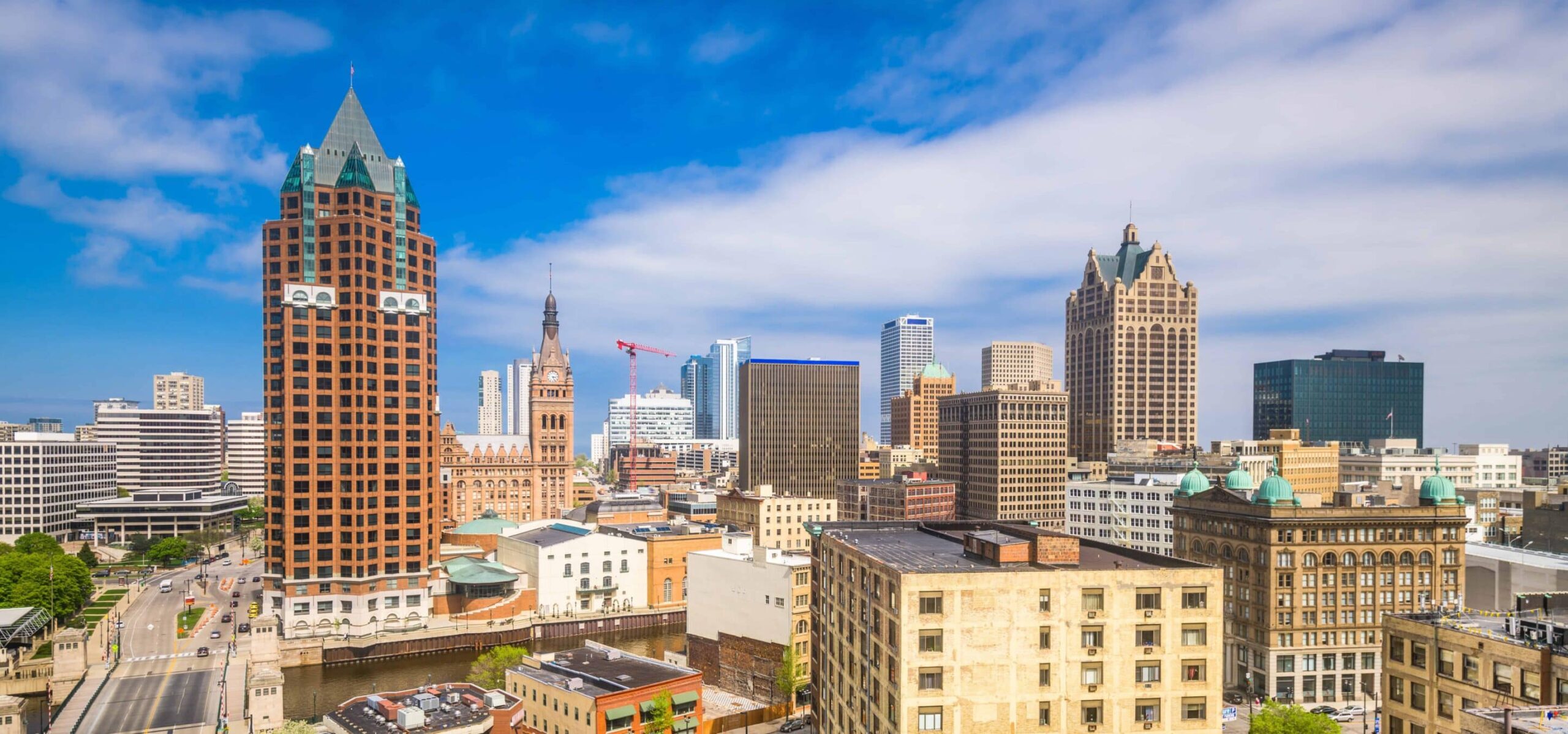 Skyline of a mid-sized city with modern and historic buildings, a red-brick landmark, cranes, high-rises under partly cloudy sky.