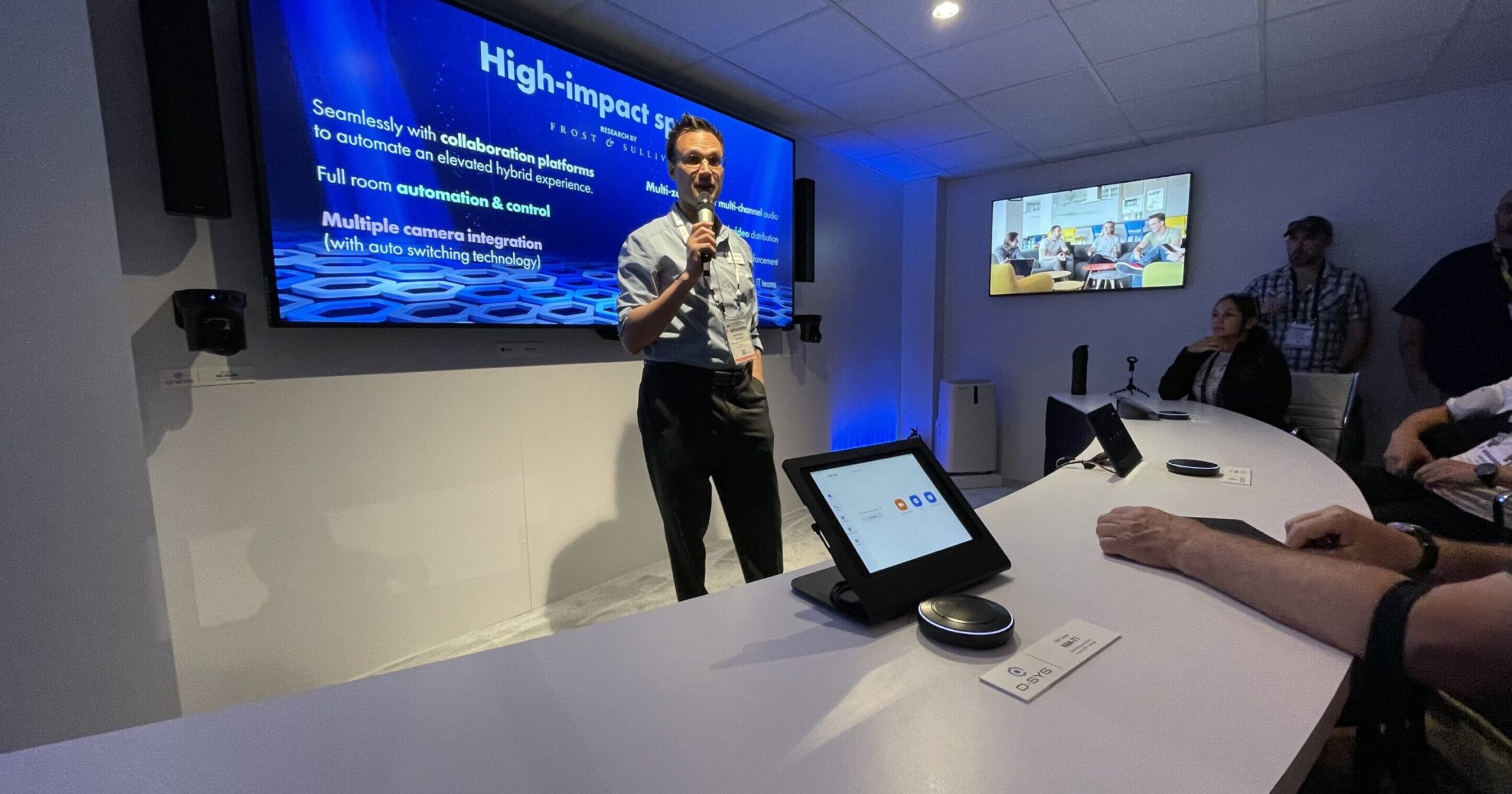 A man presents in a dim room with a curved table & screen, attendees use laptops; video conference shown.