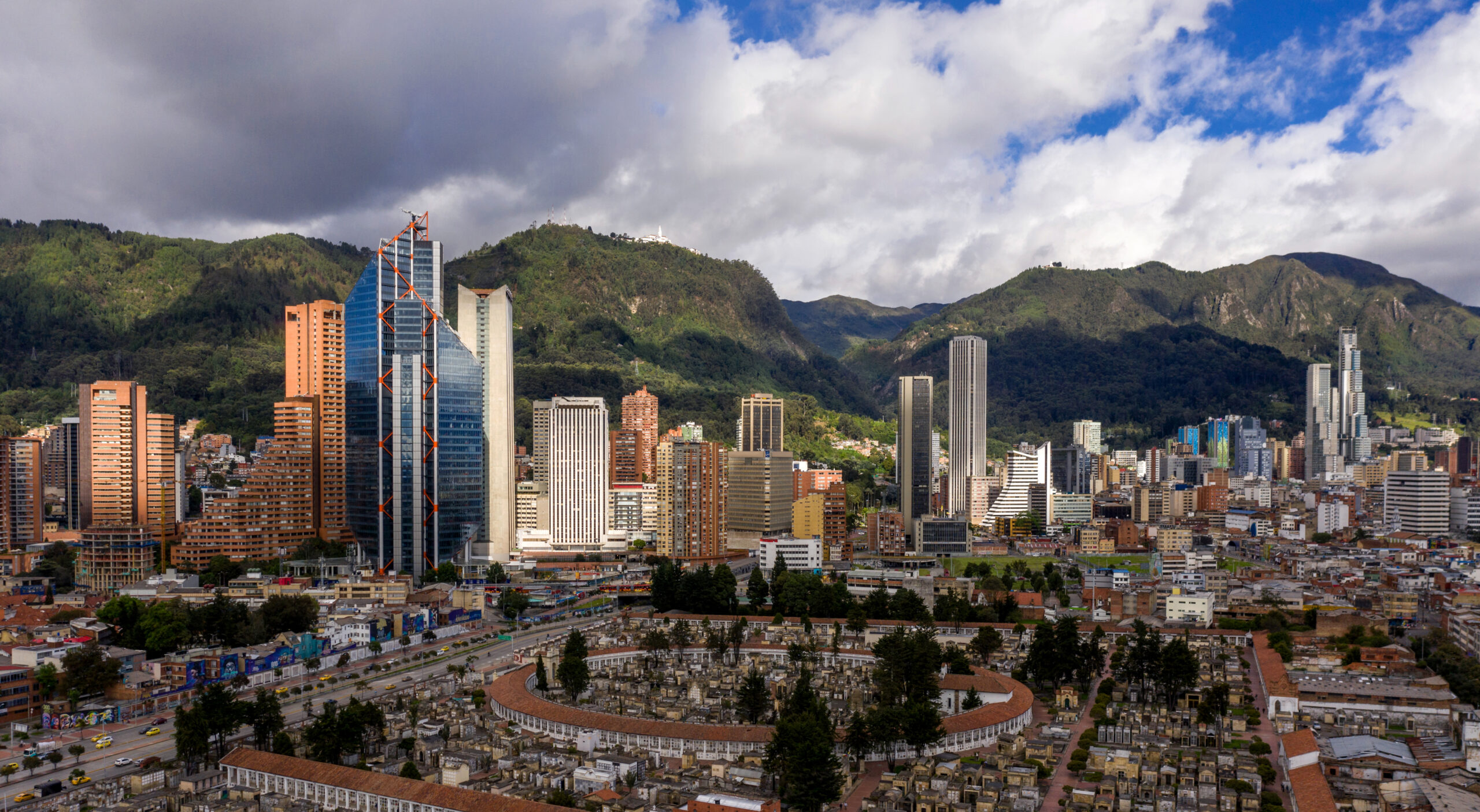 Aerial view: city skyscrapers, oval cemetery foreground, scenic mountains backdrop under a cloudy blue sky.