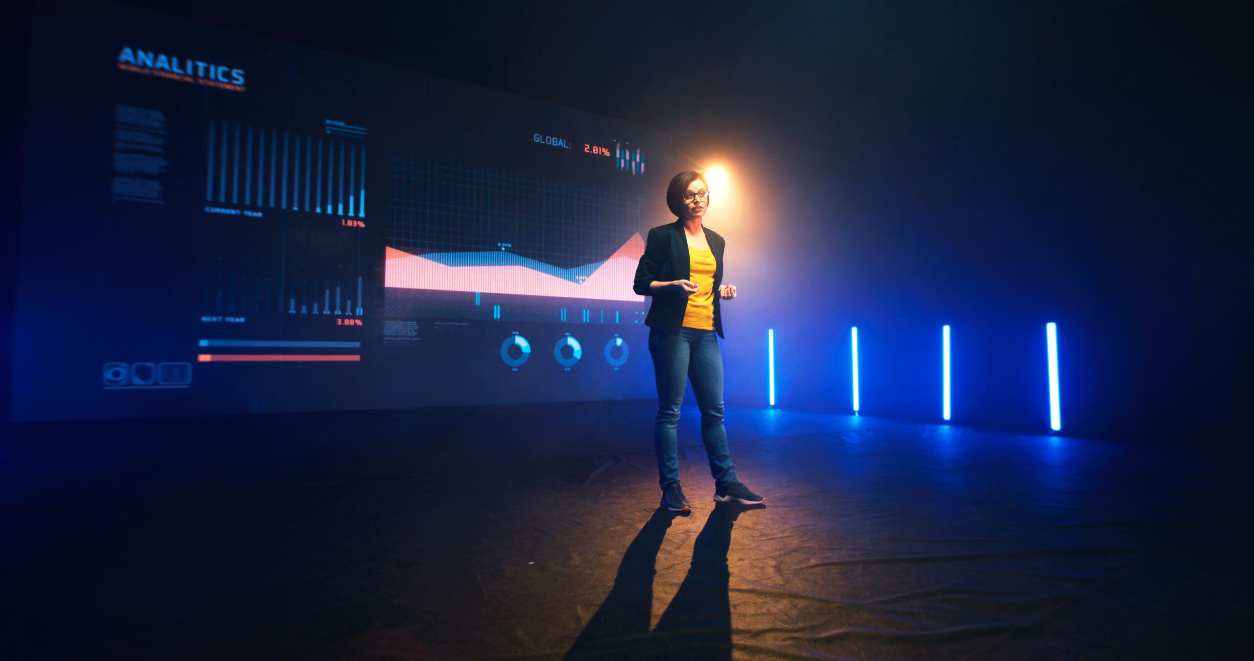 A person stands on stage presenting Analytics data on a screen, with blue lighting enhancing the dimly lit room.